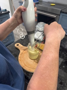 Hands using an immersion blender to blend homemade mayonnaise in a mason jar on a wooden cutting board