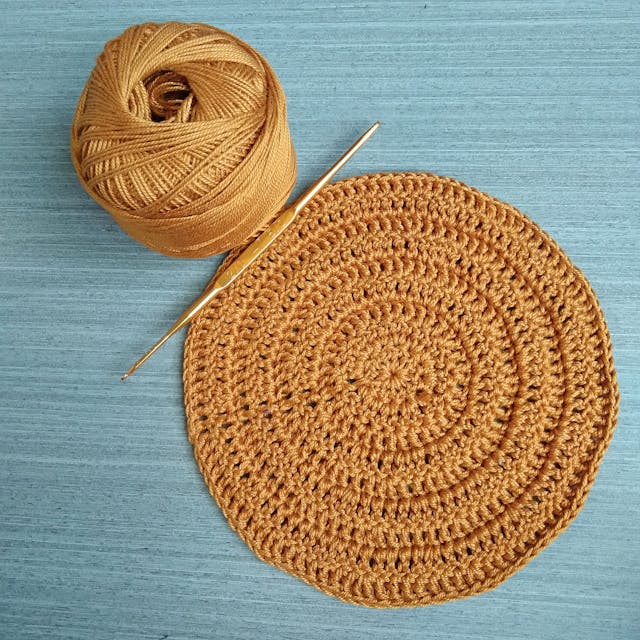 Round crochet dishcloth made with Lily Sugar'n Cream cotton yarn next to a matching skein on blue background.