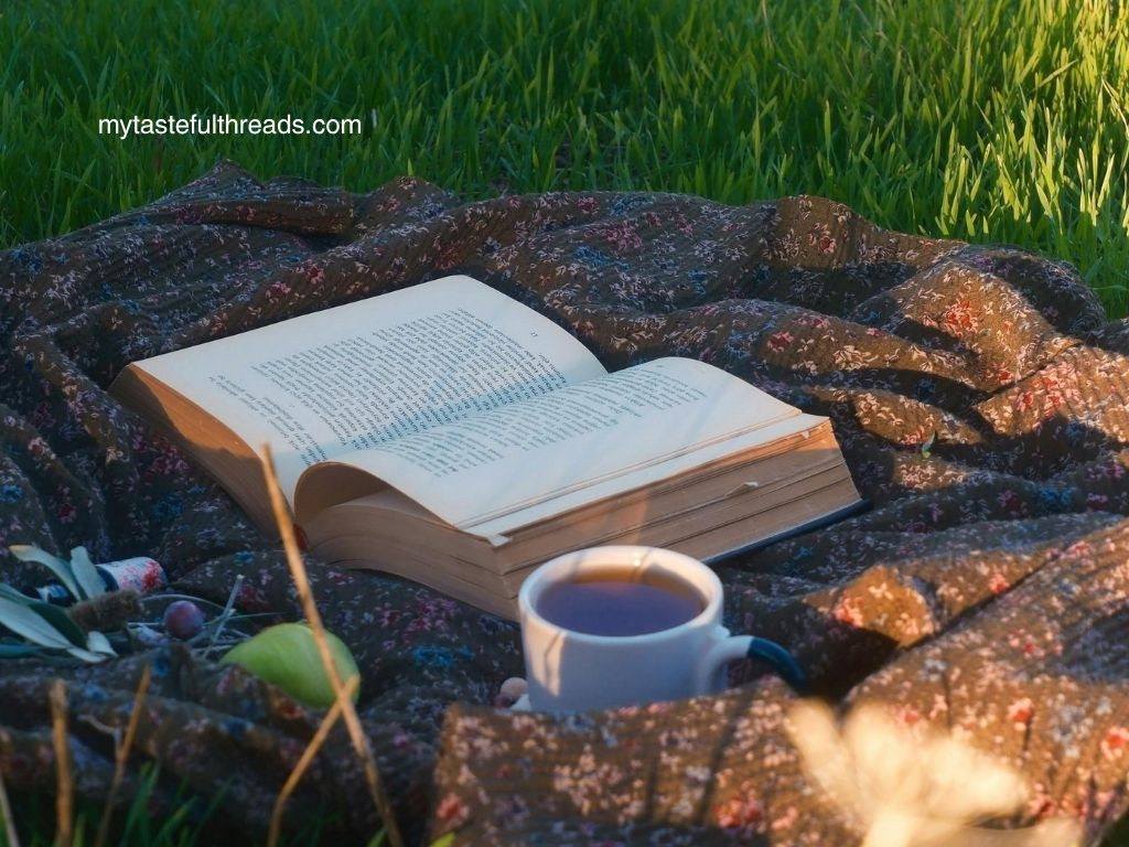 Open book and cup of tea on a floral blanket in a sunlit green meadow