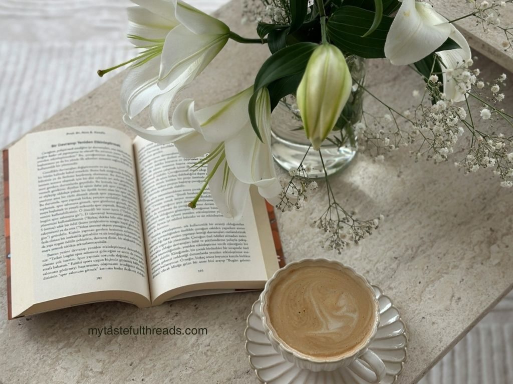 Open book on a marble surface beside a latte and a vase of white lilies and baby's breath