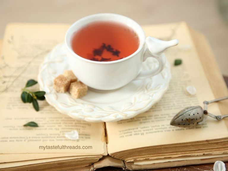 White porcelain tea cup with red tea on an open vintage book with brown sugar cubes and a tea strainer