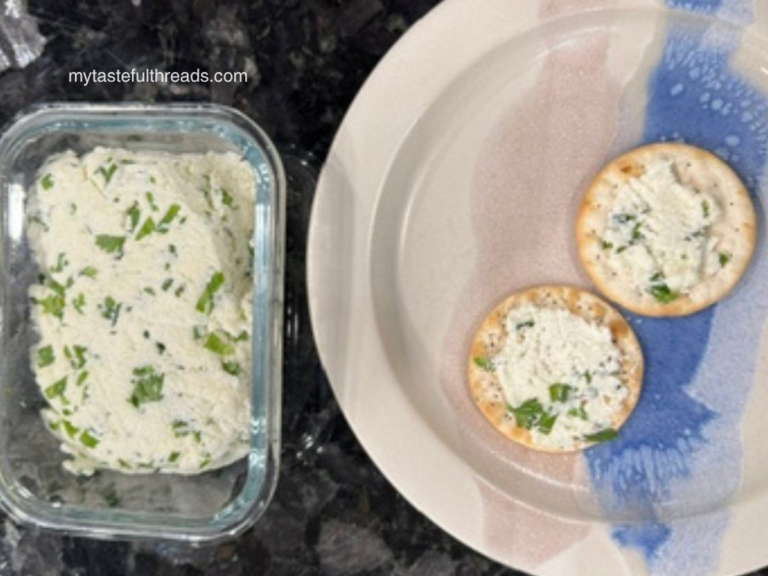 Homemade herbed goat cheese in a glass storage container next to crackers topped with fresh chèvre