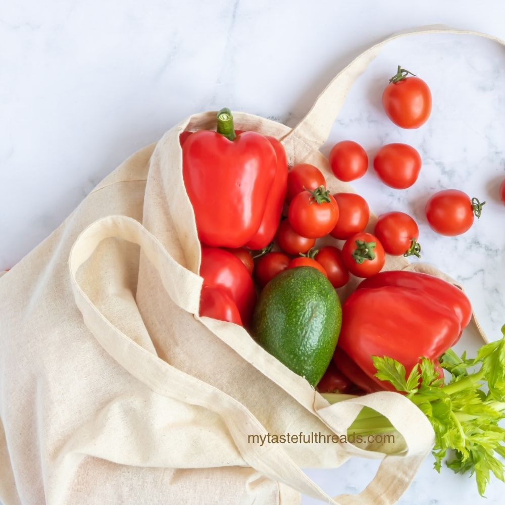 Canvas reusable grocery bag filled with peppers, tomatoes, avocado, and celery on a marble countertop